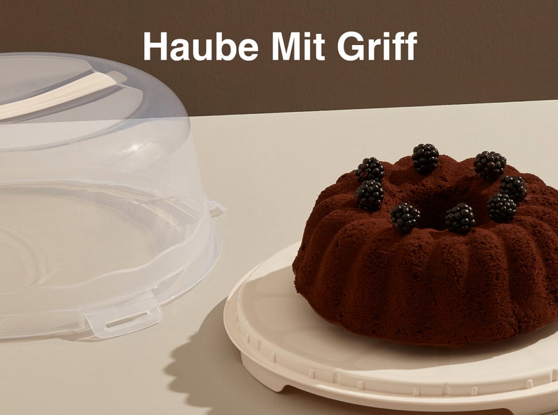 May include: A clear plastic cake dome with a handle, a white cake stand, and a chocolate cake with blackberries on top. The text "Haube Mit Griff" is written in white on a brown background.