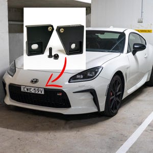 May include: Black license plate mounting brackets and hardware, displayed with a white sports car. The brackets are designed to attach to the front bumper. The car has a black grill and a license plate.