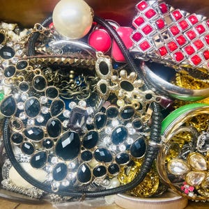 May include: A box overflowing with various jewelry pieces. Included are necklaces with black and clear stones, a large pearl, red and silver bracelets, and gold-toned pieces. A variety of styles and colors are present.