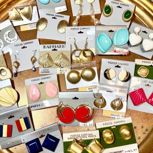 May include: A collection of vintage pierced earrings in various styles and colors, including gold, red, blue, and white. The earrings are displayed on cards, showcasing a variety of designs, from geometric shapes to pearl drops.