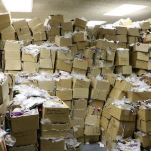 May include: A large pile of cardboard boxes, some open, filled with various items. The boxes are stacked haphazardly, creating a cluttered storage scene. Some boxes have handwritten labels. The contents are partially visible, wrapped in plastic.