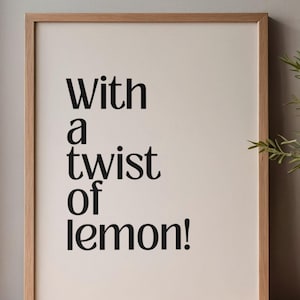 Può includere: Stampa incorniciata con la scritta "With a twist of lemon!" in nero. Il testo è centrato su uno sfondo color crema, racchiuso in una cornice di legno marrone chiaro. La stampa è esposta su una superficie di legno.