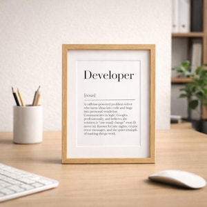Puede incluir: Un marco con la definición de "Developer". El texto describe a un solucionador de problemas impulsado por cafeína. El marco marrón claro está sobre un escritorio de madera, con teclado, ratón y portalápices.