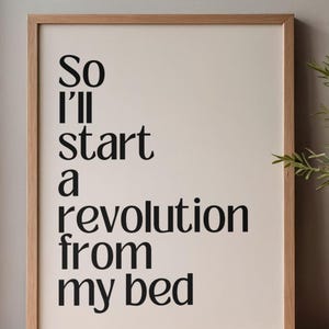 May include: A framed print with the quote "So I'll start a revolution from my bed" in black text on a cream background. The frame is made of light-colored wood. A sprig of greenery is visible on the right side.