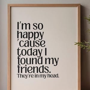 Può includere: Una stampa incorniciata con il testo "I'm so happy 'cause today I found my friends. They're in my head." Il testo nero è centrato su uno sfondo color crema, incorniciato in una cornice di legno marrone chiaro. La stampa è su una superficie di legno.