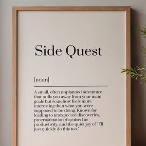 May include: A framed print with the words "Side Quest" in a bold, black font. Below, a definition describes a side quest as an unplanned adventure. The frame is a light brown color, and the background is a cream color.