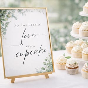 Op de afbeelding: Een goudkleurig bord met de tekst "All you need is love and a cupcake" staat op een klein ezel. Cupcakes met witte glazuur en bloemdecoraties worden gepresenteerd op een etagère, wat zorgt voor een zoete en elegante presentatie.