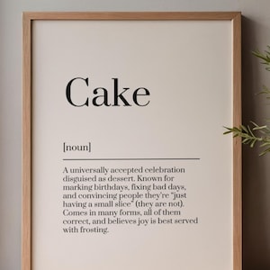 May include: A framed print with the word "Cake" in large, bold, black letters. Below, a definition describes cake as a dessert for celebrations. The frame is a light brown wood, and the background is off-white.