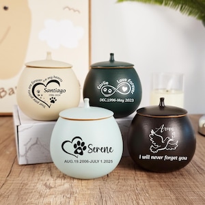 May include: Four ceramic pet urns in cream, dark green, light blue, and black. Each urn has a lid and personalized text with names, dates, and paw prints. The urns are on a wooden surface.