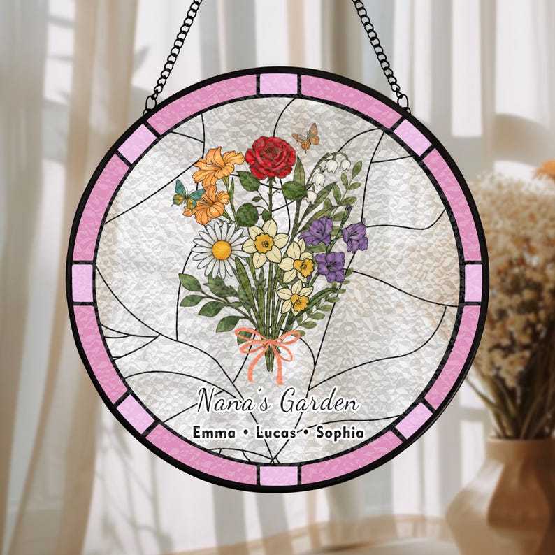 May include: Round stained glass window with a pink border and a black chain for hanging. The window features a bouquet of colorful flowers, butterflies, and the text "Nana's Garden" with three names below.