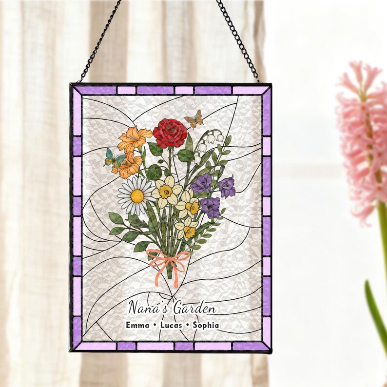 May include: Stained glass panel featuring a floral bouquet with a red rose, orange and yellow flowers, and butterflies. The panel is framed in purple and has the text "Nana's Garden" with three names below. It hangs from a black chain.