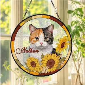 Op de afbeelding: Ronde glas-in-lood kattenportret met een lapjeskat, zonnebloemen en de naam "Nathan". De kat heeft oranje, zwarte en witte vacht. De zonnebloemen zijn geel en bruin. Het glas is omlijst met een bruine rand en een zwarte ketting om op te hangen.