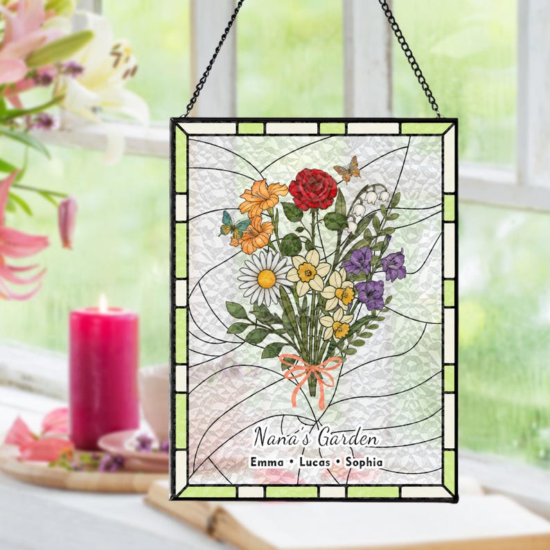 May include: Stained glass panel featuring a colorful floral bouquet with a red rose, orange flowers, and daisies. The panel is framed in black and has the text "Nana's Garden" with names below. A chain is attached for hanging.
