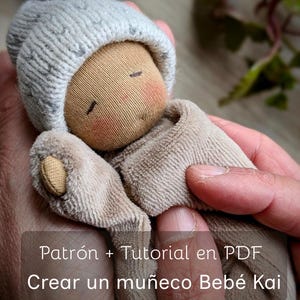 Puede incluir: Una muñeca hecha a mano con una cara de tela marrón claro, mejillas rosadas y ojos cerrados, que lleva un gorro de punto gris y un atuendo beige. La muñeca está sostenida en las manos de una persona. El texto en la imagen dice "Patrón + Tutorial en PDF Crear un muñeco Bebé Kai."