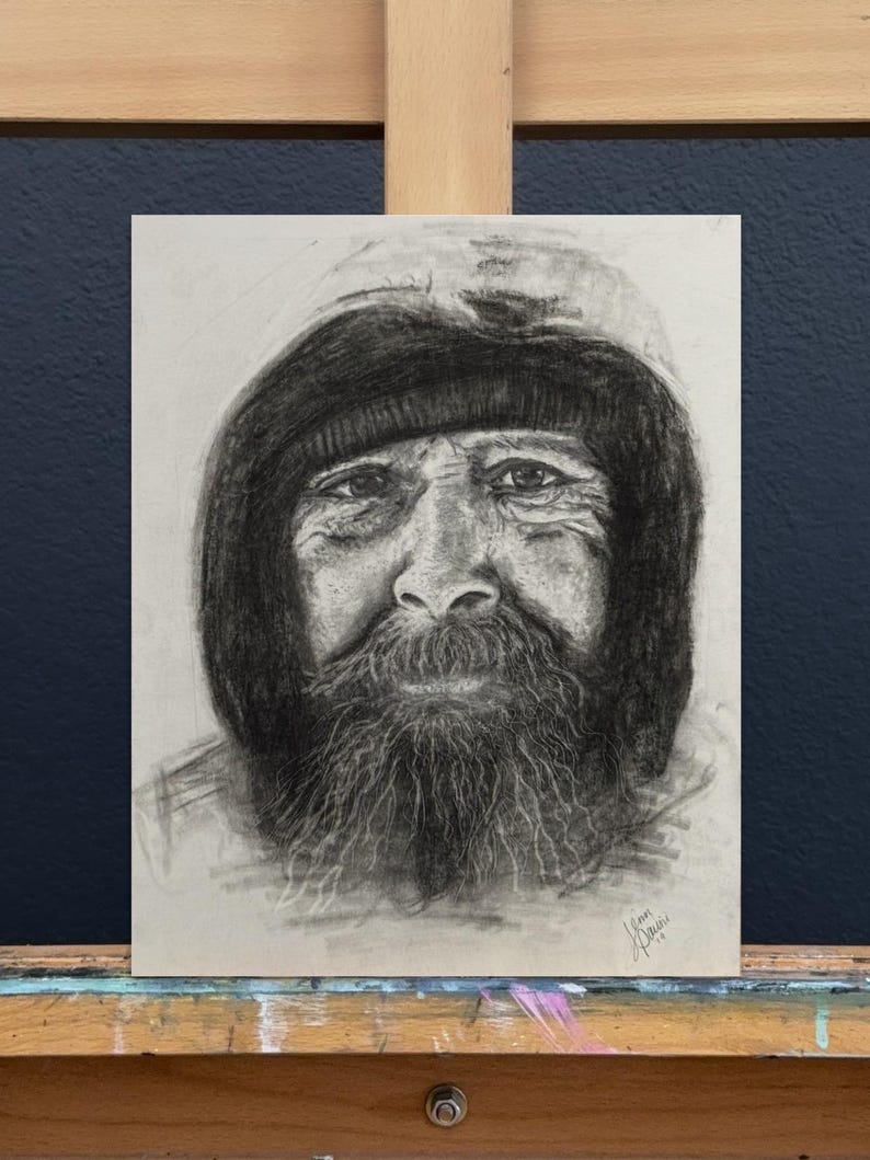 Puede incluir: Un retrato al carboncillo de una persona mayor con barba larga y prenda con capucha. La obra de arte se exhibe en un caballete, mostrando el sombreado detallado y la textura del dibujo. El rostro del sujeto es el punto focal, capturando su expresi&oacute;n.