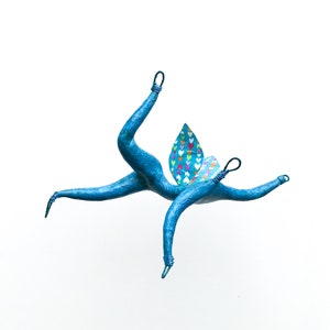 May include: A blue paper mache sculpture of a stylized butterfly with a wing made of fabric with a pattern of colorful hearts. The sculpture is suspended in mid-air.
