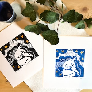 Può includere: Due stampe a linoleum blu e bianche di una donna che allatta un bambino con stelle dorate e nuvole sullo sfondo. Le stampe sono numerate 12/100 e 2/100.