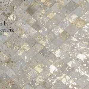 May include: A white and gold cowhide rug with a diamond pattern. The rug has a distressed look with gold accents. The rug is made of real cowhide and is a unique and stylish addition to any home.