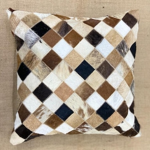 May include: A square cushion with a diamond pattern made from different shades of brown, black, and white cowhide.