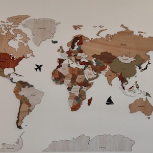 Op de afbeelding: Een houten wereldkaart met individuele landstukken in verschillende houttinten, gemonteerd op een witte muur. Inclusief zwarte vliegtuig- en bootuitsparingen en een kompasroos. De kaart is een decoratief stuk.