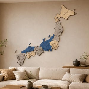 Op de afbeelding: Een grote, meerlaagse houten wandkaart van Japan met verschillende secties in natuurlijke hout-, grijze en blauwe tinten. De kaart is onderverdeeld in prefecturen met gegraveerde labels en is bevestigd aan een muur boven een beige bank.