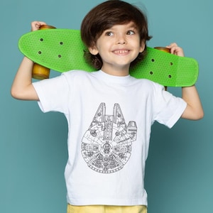 May include: A white t-shirt featuring a detailed black and white illustration of the Millennium Falcon. The shirt is worn by a child holding a green skateboard. The background is a solid teal color.