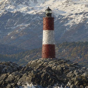 May include: A cylindrical lighthouse featuring horizontal red and white stripes, topped with a dark lantern room. The structure stands prominently on a dark, jagged rocky outcrop, set against a backdrop of snow-capped mountains and dense forest.