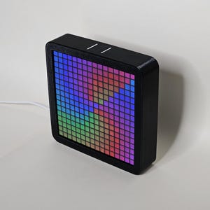 Könnte beinhalten: Ein schwarzes quadratisches Gerät mit einem bunten Pixeldisplay, das ein Regenbogenmuster zeigt. Das Gerät ist mit einem weißen Kabel angeschlossen.