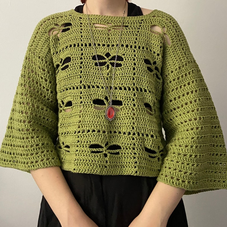 Puede incluir: Un su&eacute;ter verde de punto de ganchillo corto con un collar de cadena plateada y un colgante de piedra roja.
