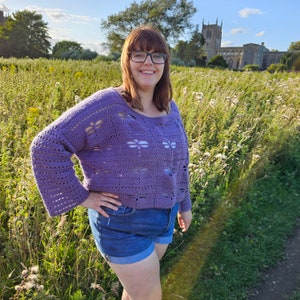 Puede incluir: Una mujer con un su&eacute;ter de crochet morado con un dise&ntilde;o de lib&eacute;lula y pantalones cortos de mezclilla azul. Est&aacute; de pie en un campo de hierba alta.