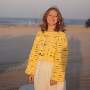 Puede incluir: Una mujer lleva un su&eacute;ter de crochet amarillo con un patr&oacute;n de lib&eacute;lula. El su&eacute;ter es corto y tiene mangas largas. Est&aacute; de pie en una playa.