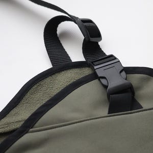 Pu&ograve; includere: Primo piano di un oggetto in tessuto verde con finiture e cinghie nere. &Egrave; visibile una fibbia in plastica nera, insieme a una cinghia nera. L'oggetto ha una morbida fodera interna strutturata. Lo sfondo &egrave; bianco.