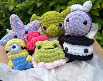 Op de afbeelding: Een verzameling kleine, handgemaakte gehaakte knuffels, waaronder een veelkleurige zeester, een geel-blauw figuurtje, een roze octopus, een groen wezen met grote oren, een lichtgroene kikker met een roze basis en een zwart-wit koekje.