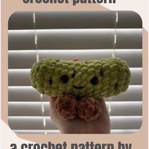 Può includere: Un modello all'uncinetto di sottaceto con il testo "pickle crochet pattern" e "a crochet pattern by the crochet store". Il sottaceto è verde con occhi neri e piedi marroni. Lo sfondo è una finestra con le persiane.