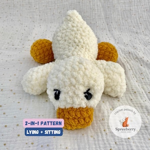 May include: A white crocheted duck plush toy with yellow crocheted accents. The duck is lying down with its head turned to the side. The toy is a 2-in-1 pattern, meaning it can be made to lie down or sit up. The pattern is by Spreeberry Creative Studio.