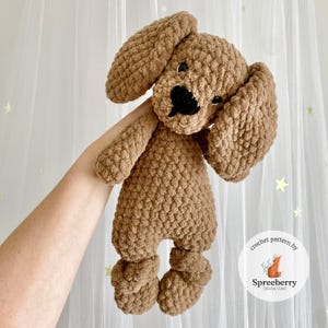 Puede incluir: Un juguete de peluche de perro de crochet marrón con ojos negros y nariz negra. El perro está siendo sostenido por una mano.