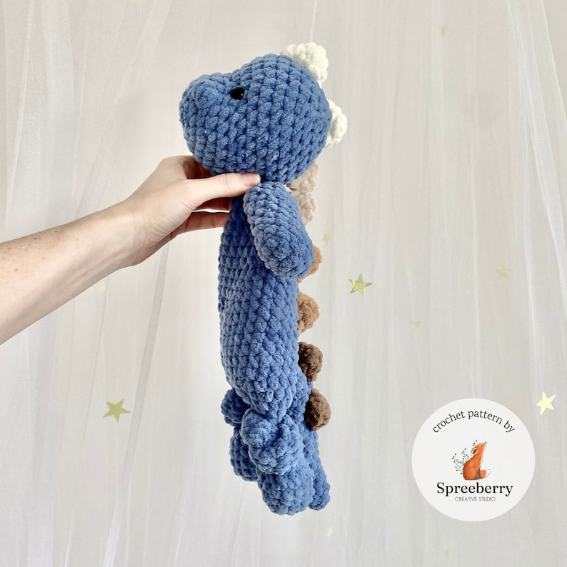 May include: A blue crocheted dinosaur plush toy with brown and white accents. The toy is being held by a hand. The image includes the text "crochet pattern by Spreeberry Creative Studio".