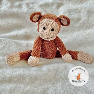 Könnte beinhalten: Ein braunes Häkel-Affenspielzeug mit weißem Gesicht und weißem Bauch. Der Affe sitzt auf einer weißen, flauschigen Decke. Der Text "crochet pattern by Spreeberry Creative Studio" befindet sich in einem Kreis unter dem Affen.