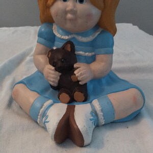 May include: A ceramic figurine of a girl with blonde pigtails wearing a blue dress and holding a brown teddy bear.