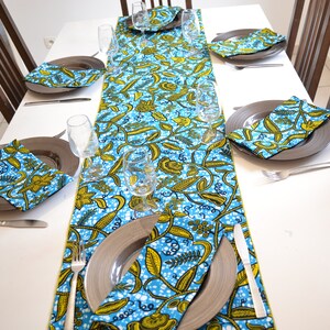 May include: A blue and yellow patterned table runner with matching napkins. The runner and napkins have a floral design with white accents. The runner is laid out on a white table with silver plates and silverware.