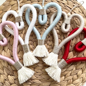 May include: Six handmade heart-shaped ornaments with a tassel. The ornaments are made of yarn in various colours, including pink, white, light blue, and red. Each ornament has a gold accent.