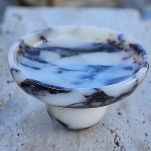 May include: A white marble bowl with black veining, featuring a pedestal base. The bowl is perfect for serving snacks or holding small items.
