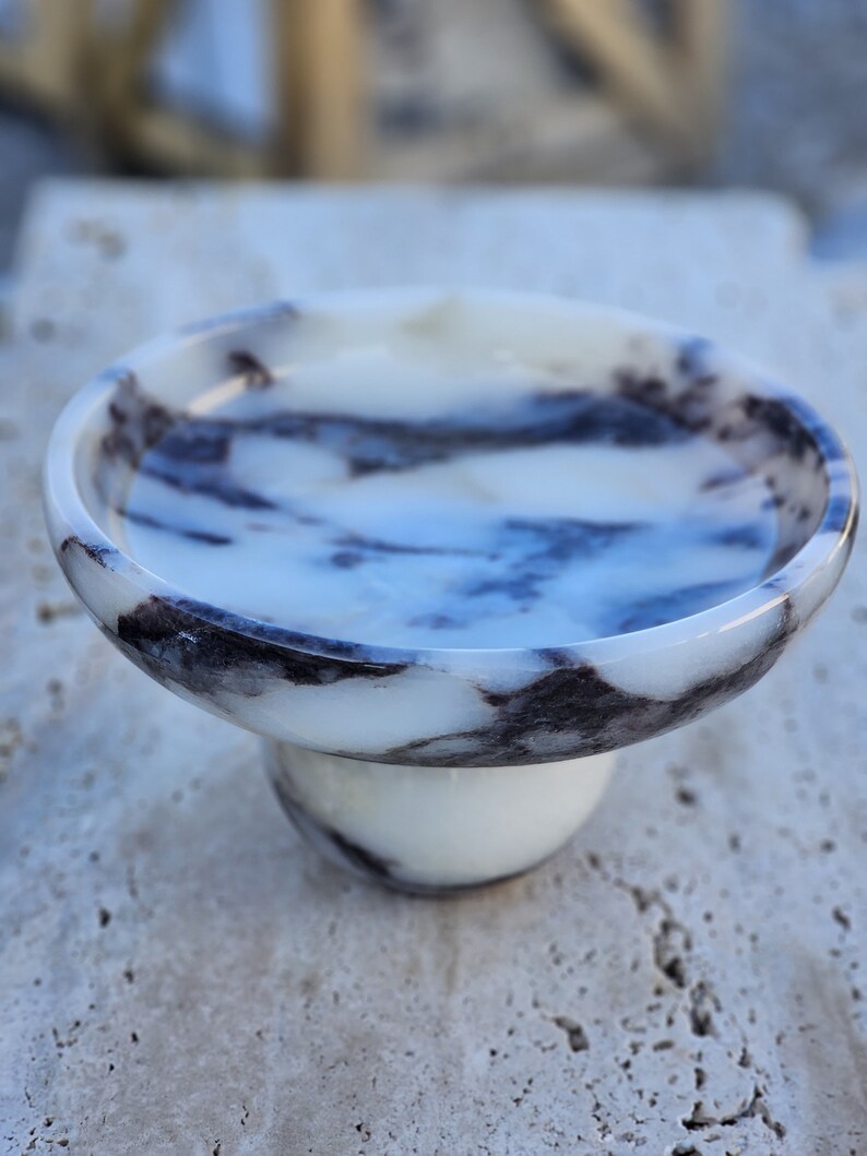 May include: A white marble bowl with black veining, featuring a pedestal base. The bowl is perfect for serving snacks or holding small items.