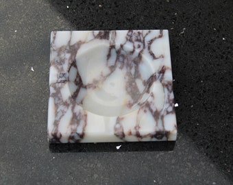 May include: Square ashtray made of white marble with brown veining. The ashtray has a circular indentation in the center for holding cigarettes or cigars. The marble has a polished finish, and the ashtray is set on a dark surface.