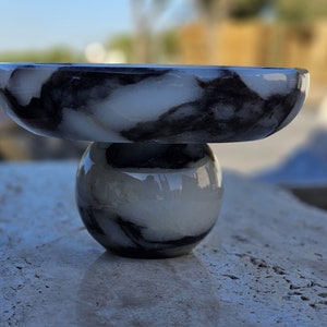 May include: A white marble bowl with black marbling sits on top of a white marble sphere with black marbling. The bowl and sphere are both made of polished marble.