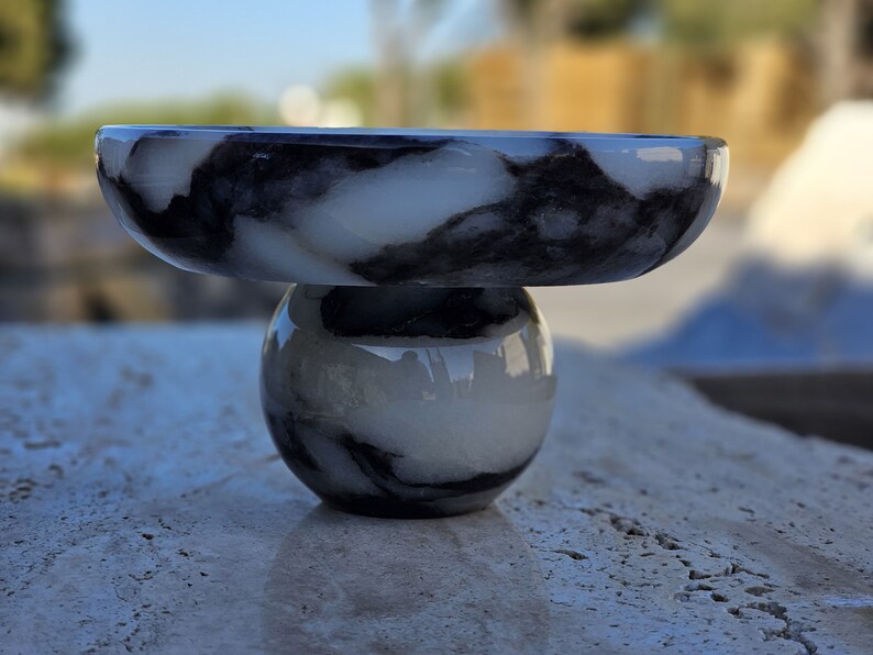 May include: A white marble bowl with black marbling sits on top of a white marble sphere with black marbling. The bowl and sphere are both made of polished marble.