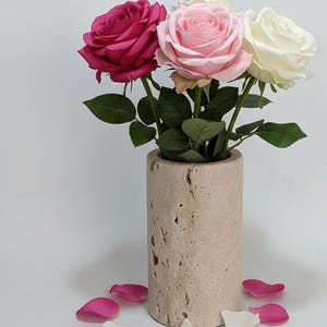 Könnte beinhalten: Ein Arrangement mit drei Rosen in einem beigefarbenen Steingut-Vase. Die Rosen sind magenta, rosa und weiß. Streuende Blütenblätter in passenden Farben umgeben die Vase und schaffen eine romantische Präsentation.