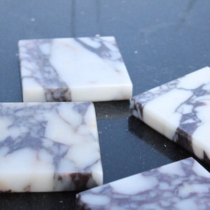 May include: Four square marble coasters with a white background and grey and purple veining. The coasters are on a black surface.