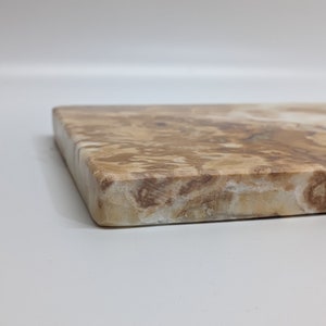 May include: A rectangular chopping board with a natural stone effect. The board has a mix of tan, brown, and cream hues, with a polished finish. The edges are gently rounded, and the design is simple and elegant.