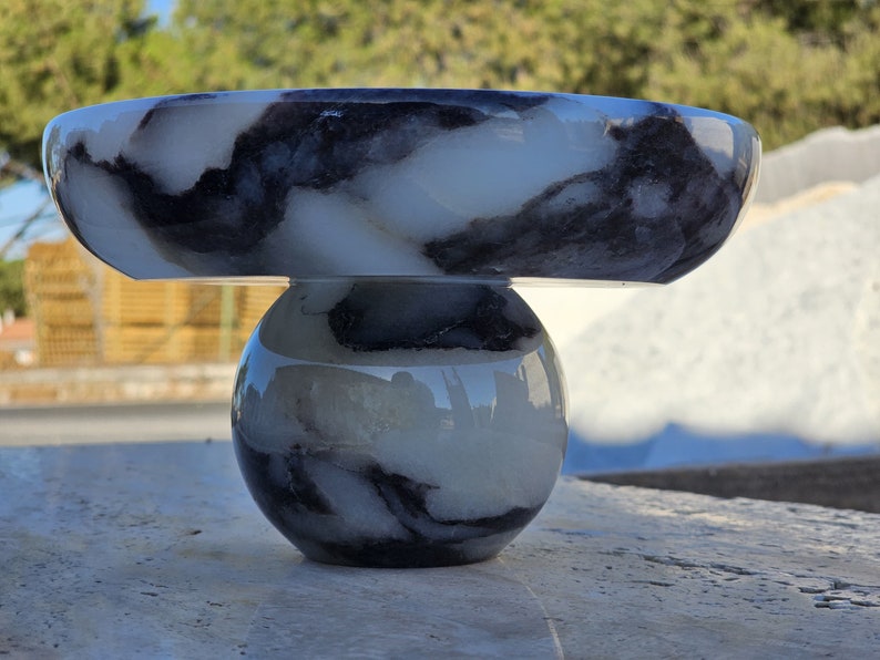 May include: A white and black marble bowl with a round base. The bowl is made of alabaster and has a unique marbled pattern.
